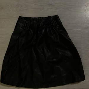 Svart kort barn kjol  i skinnimitatin  - Snygg svart kortkjol i skinnimitation   med resår i midjan. Kjolen har en enkel och stilren design som ger en bra vibe till din outfit. Perfekt att matcha med en vit tröja.                                                     Storlek 6Y/ 6 åringar 