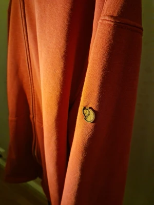 Rostfärgad hoodie från Fjällräven - Säljer en snygg rostfärgad hoodie från Fjällräven med dragkedja framtill och klassisk logga på ärmen. Hoodien har huva med vita snören och kängurufickor. Tillverkad i mjukt bomullsmaterial som är skönt att ha på sig.