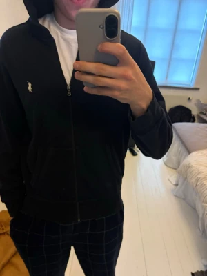 Marinblå hoodie från Polo Ralph Lauren - Säljer en marinblå hoodie från Polo Ralph Lauren med dragkedja och klassiska loggan broderad på bröstet. Hoodien har huva och ribbade muddar. Perfekt för en chill och stilren look. Materialet känns mjukt och skönt mot huden. 