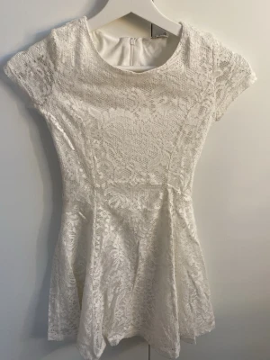 Vit spetsklänning med kort ärm - Söt vit kortklänning med spetsdetaljer och rund halsringning. Klänningen har korta ärmar, figurnära passform upptill och utställd kjol. Dragkedja bak för enkel påklädning. Perfekt för sommarens alla tillfällen.