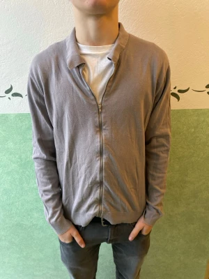 Ljusgrå ziptröja från Riley - Ljusgrå ziptröja från Riley med krage och hel dragkedja framtill. Tröjan har långa ärmar och ribbade muddar vid ärmslut och nederkant. Perfekt för en avslappnad och stilren look.