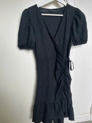 Svart omlottklänning med puffärm - Svart kort omlottklänning med puffärmar och volangkant nedtill. Klänningen har knyt i midjan och v-ringad hals. Endast använd 1 gång!