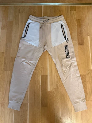 Beige mjukisbyxor från Hoodrich - Snygga beige mjukisbyxor från Hoodrich med vita paneler framtill och svarta dragkedjor på fickorna. Byxorna har resår i midjan med snörning, muddar vid bensluten och tryckt logga på ena benet samt bakficka med logga. Perfekta för en chill och trendig look.