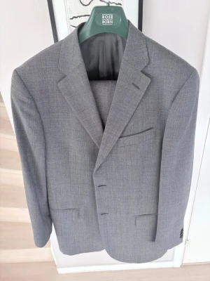 Grå kavaj från Rose & Born - Stilren och exklusiv grå kostymset från lyxmärket Rose & Born. Kavajen är i klassisk modell med två knappar, spetsiga slag och flera fickor. Tunt rutmönster i tyget och helfodrad insida. Perfekt för en dressad look och passar till både skjorta och t-shirt. Nypris är runt 15.000-20.000 kr. 
