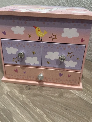 Smyckeskrin med fågel och moln - Sött smyckeskrin i rosa och lila med motiv av fågel, moln, stjärnor och hjärtan. Skrinet har tre lådor och ett fack med spegel samt en snurrande enhörning. Perfekt för att förvara smycken och småaccessoarer. Material: trä och textilklädd insida.