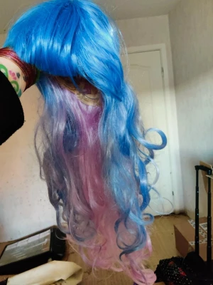 Blå och rosa lockig cosplayperuk - Snygg lång peruk med intensivt blått upptill som övergår i pastellrosa i längderna. Peruken har stora, mjuka lockar och är perfekt för cosplay eller maskerad. Tillverkad i syntetmaterial och har en naturlig glans.