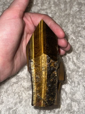 Stor polerad tigeröga kristallspets - Säljer en stor, polerad tigeröga kristall med spetsig form. Kristallen har vackra gyllene och bruna band i materialet och är slipad på ena sidan medan andra sidan är mer rå. Perfekt som dekoration eller samlarobjekt.