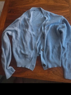 Ljusblå ribbad kofta med knappar - Snygg ljusblå kofta med ribbad struktur och knäppning framtill. Koftan har långa ärmar och breda muddar vid ärmslut och nederkant. Perfekt att slänga över en t-shirt för en chill look.