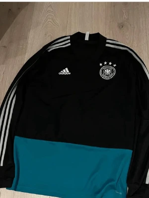 Adidas Tyskland långärmad träningströja - Snygg långärmad träningströja från Adidas med Tysklands landslagslogga och fyra stjärnor på bröstet. Tröjan är svart med gråa ränder på ärmarna och har en turkos nederdel. Tillverkad i lätt och funktionellt material, perfekt för fotbollsträning.