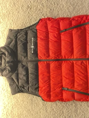 Röd och svart dunväst från Sailracing - Snygg dunväst från Sailracing i rött och svart med hög krage och två dragkedjeförsedda fickor framtill. Västen har quiltad design och logga på bröstet samt nacken. Perfekt för lager-på-lager och håller dig varm under kyliga dagar. Hör av dig vid minsta fundering mvh Artur !