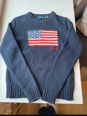 Blå stickad tröja Polo Ralph Lauren - Mörkblå stickad tröja från Polo Ralph Lauren med amerikansk flagga i rött, vitt och blått på bröstet samt RL-broderi. Tröjan har rund halsringning, ribbade muddar och är gjord i mjukt material. Perfekt för dig som gillar klassisk preppy stil.