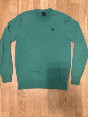 Grön stickad tröja från Peak Performance - Snygg grön stickad tröja från Peak Performance med rund halsringning och diskret svart logga på bröstet. Tröjan har långa ärmar och ribbade muddar vid ärmslut och nederkant. Perfekt för lager på lager och enkel att matcha.