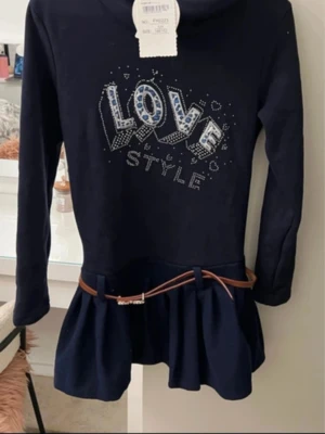 Marinblå klänning med text och bälte - Mörkblå klänning med lång ärm och texten 'LOVE STYLE' i glittriga stenar på bröstet. Kjoldelen har volang och midjan pryds av ett smalt brunt bälte. Perfekt för dig som gillar en trendig och ungdomlig look.