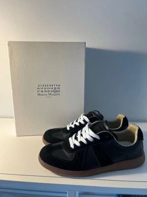 Maison Margiela svarta sneakers - Maison Margiela sneakers i svart läder och mocka med vita skosnören och brun gummisula. Klassisk låg modell med stilren design och diskreta detaljer.(kan ej garantera äkthet pga att jag inte har nått kvitto )Perfekta för dig som gillar minimalistisk och trendig streetwear.