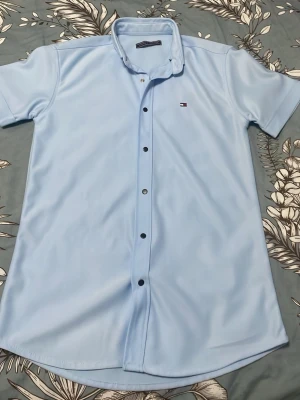 Ljusblå kortärmad skjorta Tommy Hilfiger - Snygg ljusblå skjorta från Tommy Hilfiger med korta ärmar och klassisk krage. Skjortan har knappar framtill och den ikoniska loggan broderad på bröstet. Perfekt för en clean och stilren look.