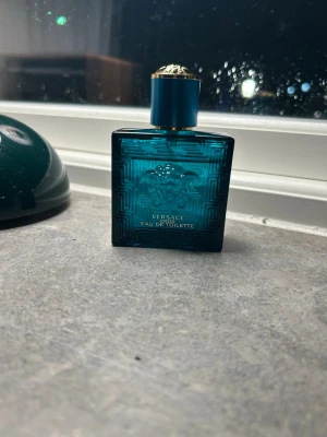 Versace Eau de Toilette blå flaska - Snygg parfym från Versace i en fyrkantig blå glasflaska med guldiga detaljer och mönstrad yta. Flaskan har en stilren design med Versaces ikoniska Medusa-logga på framsidan. Perfekt för dig som gillar exklusiva dofter och vill ha en lyxig parfym i samlingen.