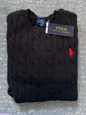 Svart kabelstickad tröja Polo Ralph Lauren - Kabelstickad svart tröja från Polo Ralph Lauren med rund halsringning och den klassiska röda loggan broderad på bröstet. Tröjan har långa ärmar och ribbade muddar vid ärmslut och nederkant. Perfekt för dig som gillar stilrena och tidlösa plagg. Tröjan är helt ny med prislapp. Storlek M. Pris kan diskuteras.