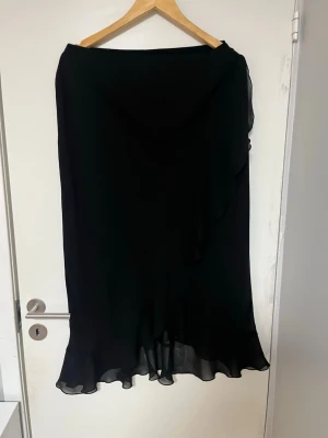 Svart långkjol med volangkant - Säljer en svart långkjol med volangkant nedtill. Kjolen har en mjuk och flytande form som ger en elegant siluett. Perfekt för dig som gillar stilrena plagg med en twist. Passar till många olika looks! Storlek 46 men insydd så den är ca storlek 44.