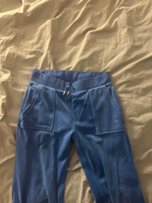Blå velour mjukisbyxor Juicy Couture - Säljer ett par blå mjukisbyxor från Juicy Couture i mjuk velour. Byxorna har bred resår i midjan med dragsko, stora fickor fram och bakfickor med knappar. Perfekta för en chill och avslappnad stil.