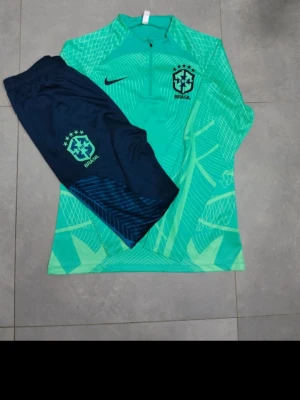 Nike Brasil träningsset grön/blå - Snyggt träningsset från Nike med Brasil-logga. Tröjan är grön med grafiskt mönster, halv dragkedja och lång ärm. Byxorna är mörkblå med ljusgrön Brasil-märkning och diskret mönster på benen. Perfekt för fotbollsträning eller gymmet.