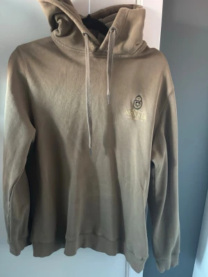 Olivgrön hoodie från Morris - Snygg olivgrön hoodie från Morris med broderad logga på bröstet. Klassisk modell med huva och dragsnören samt ribbade muddar vid ärmslut och nederkant. Tillverkad i mjukt bomullsmaterial som känns skönt mot huden.