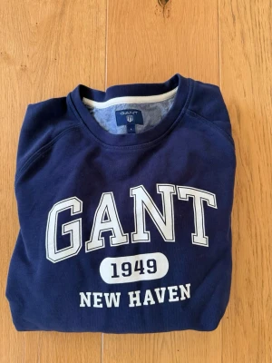 Marinblå sweatshirt från GANT - Klassisk marinblå sweatshirt från GANT med tryck 'GANT 1949 NEW HAVEN' på bröstet. Tröjan har rund halsringning, långa ärmar och ribbade muddar vid ärmslut och nederkant. Perfekt för en avslappnad och schysst stil.