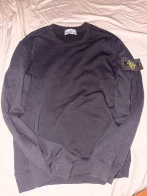Stone island tröja - Svart sweatshirt från Stone Island med ikonisk kompasspatch på vänster ärm. Tröjan har rund hals, ribbade muddar och är gjord i mjuk bomull. Enkel och clean design som passar perfekt till streetwear-stilen.
