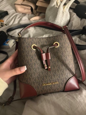 Brun bucket bag från Michael Kors - Snygg brun bucket bag från Michael Kors med monogrammönster och detaljer i mörkrött läder. Väskan har guldfärgade metalldetaljer, justerbar axelrem och dragskostängning. Perfekt storlek för det viktigaste och ger en trendig touch till din outfit.
