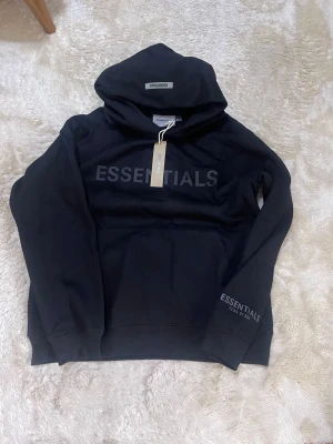 Svart Essentials Fear of God hoodie - Säljer en svart hoodie från Essentials Fear of God med stor logga på bröstet och mindre tryck på ärmen. Hoodien har huva, känguruficka och ribbade muddar. Tillverkad i mjukt bomullsmaterial, perfekt för en avslappnad streetwear-look.