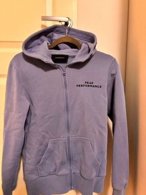 Blå hoodie Peak Performance - Snygg blå hoodie från Peak Performance med dragkedja framtill och två stora fickor. Klassisk huva och ribbade muddar vid ärmslut och nederkant. Diskret logga på bröstet. Perfekt för en avslappnad och sportig stil.