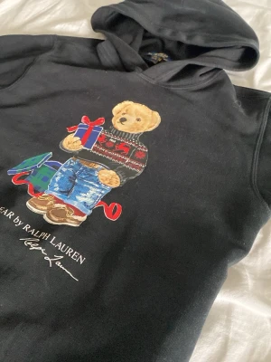 Svart hoodie Polo Bear Ralph Lauren - Svart hoodie från Ralph Lauren med ikoniska Polo Bear-trycket framtill. Björnen har på sig en stickad tröja och jeans. Hoodien har huva och ribbade muddar. Perfekt för dig som gillar grisch stilen. Köptes för 1500 mitt pris är 672