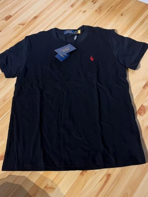 Svart t-shirt från Polo Ralph Lauren - Klassisk svart t-shirt från Polo Ralph Lauren med röd broderad logga på bröstet. Tillverkad i mjuk bomull och har en normal passform med korta ärmar och rund halsringning. Perfekt basplagg för en clean och stilren look.