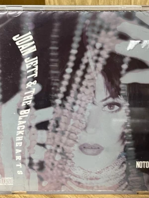 CD Joan Jett & The Blackhearts - CD-skiva med Joan Jett & The Blackhearts. Omslaget har ett svartvitt foto med pärlor och artistens ansikte i fokus. Baksidan visar bandmedlemmarna och låtlista. Släppt av Sony Music 1991. Perfekt för dig som gillar rock och samlar på klassiska album.