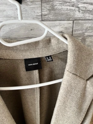 Beige kappa från Vero Moda - Stilren beige kappa från Vero Moda med klassisk krage och knytskärp i midjan. Kappan har en rak passform.