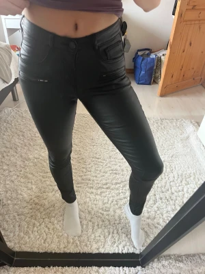 Svarta skinnbyxor  - Säljer ett par svarta leggings i skinn-look med hög midja och smal passform. De har fickdetaljer fram och bak samt knapp och dragkedja framtill.