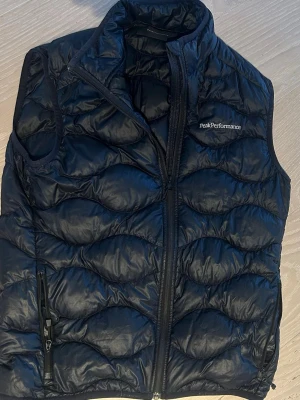 Svart dunväst från Peak Performance - Snygg svart dunväst från Peak Performance med quiltat mönster och dragkedja framtill. Västen har en hög krage och en praktisk ficka med dragkedja på sidan. Perfekt för lager på lager och håller dig varm under kyliga dagar.