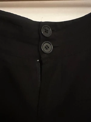 Svarta utsvängda kostymbyxor - Snygga svarta kostymbyxor med utsvängda ben och hög midja. Byxorna har två knappar framtill och hällor för skärp. Perfekta för en stilren look och passar till många olika outfits.