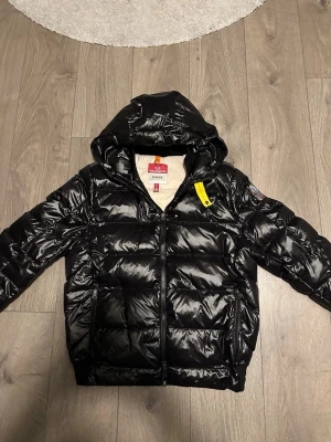 Parajumper sheen svart - Snygg svart dunjacka från Parajumpers med glansig finish, huva och dragkedja framtill. Jackan har en gul detalj vid halsen och Parajumpers-logga på ärmen. Perfekt för kalla vinterdagar och riktigt trendig look.