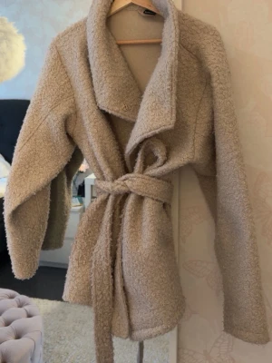 Beige teddyjacka med knytskärp - Mysig beige teddyjacka med bred krage och knytskärp i midjan. Jackan har en oversized passform och är gjord i fluffigt teddy-material som ger en trendig look. Perfekt för att hålla värmen under kyliga dagar och passar till många olika outfits.