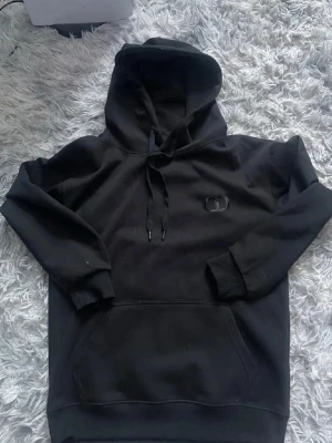 Svart hoodie  - Säljer en svart hoodie med broderad logga på bröstet. Hoodien har klassisk känguruficka, dragsko i huvan och ribbade muddar vid ärmar och nederkant. Perfekt för dig som gillar enkel och stilren streetwear.
