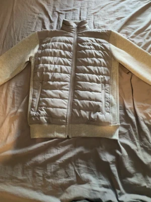 Ljusgrå Tommy Hilfiger cardigan med dragkedja  - Snygg ljusgrå cardigan från Tommy Hilfiger med quiltad front och mjuka ärmar i sweatshirtmaterial. Jackan har hög krage, två fickor med dragkedja och ribbade muddar. Perfekt för kalla vinterdagar och ger en sportig look.