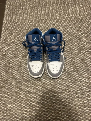 Nike Air Jordan 1 Mid grå/blå - Snygga Nike Air Jordan 1 Mid sneakers i vitt, grått och blått.Perfekta för dig som gillar streetwear och ikonisk design. Andvända fåtal gånger men har mycket kvar att ge! Nypris: 1500kr. Hör av er vid funderingar✌🏼
