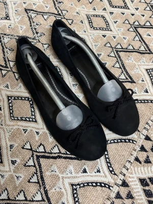 Svarta ballerinaskor med rosett - Stilrena svarta ballerinaskor med rund tå och dekorativ rosett framtill. Skorna har en klassisk och enkel design som passar till många olika outfits. Materialet är i mocka för en mjuk och matt finish.