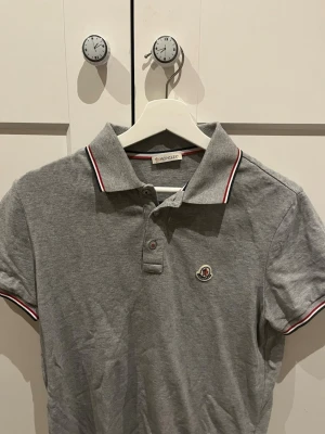 Grå Moncler piké  - Grå piké från Moncler med klassisk krage, två knappar och rödvita detaljer på krage och ärmslut. Ikonisk Moncler-logga på bröstet. Tillverkad i mjuk bomull för en clean och stilren look.