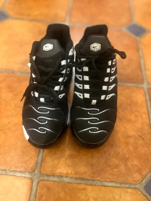 Nike Air Max Plus TN skor  - Nike Air Max Plus sneakers i svart och vitt med ikoniska vågmönster på sidan och synlig Air-dämpning i sulan. Skorna har mesh- och syntetmaterial, klassisk Swoosh-logga och snörning. Perfekta för dig som gillar streetwear och sportig stil.