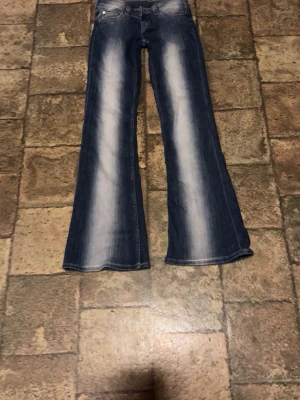 Blå bootcut jeans med slitningar - Säljer ett par blå bootcut jeans ljusa partier framtill och baktill. Jeansen har en klassisk femficksmodell och ljusare partier längs benen för en cool vintage-look. Perfekta för dig som gillar retrostil och vill ha ett par jeans som sticker ut.