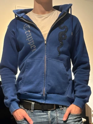Blå hoodie med orm och text - Blå zip-hoodie med huva och fickor framtill. På ena sidan av bröstet finns en orm i svart/vitt tryck och på andra sidan står det 'Chrome' i glittriga bokstäver. Tillverkad i mjukt sweatshirtmaterial, perfekt för en avslappnad stil.