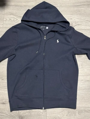 Mörkblå hoodie från Polo Ralph Lauren - Snygg mörkblå hoodie med dragkedja från Polo Ralph Lauren. Klassisk design med vit broderad logga på bröstet, två stora fickor framtill och justerbar huva. Tillverkad i mjuk bomullsblandning som är skön att bära. Perfekt för en avslappnad och stilren look.