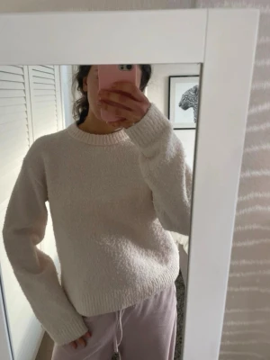 Ljusbeige fluffig stickad tröja - Mysig ljusbeige stickad tröja med rund hals och långa ärmar. Tröjan har en fluffig, mjuk struktur och ribbade muddar vid ärmslut och nederkant. Perfekt för kyliga dagar och enkel att matcha med olika stilar.