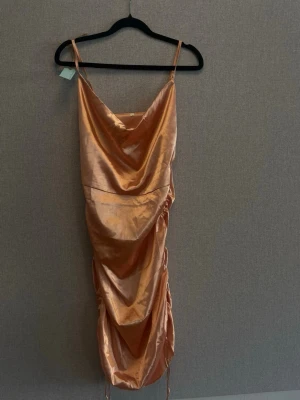 Champagnefärgad satin klänning - Snygg kort klänning i champagnefärgad satin med draperad framsida och tunna axelband. Klänningen har en omlottliknande effekt och en glansig finish som ger en lyxig känsla. Perfekt för dig som vill sticka ut.